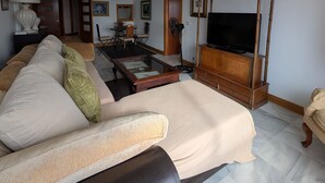 A living room with a sofa, a coffee table, a TV on a wooden stand, and a dining area in the background. - 293 - Apartamento El Puerto (Tarifa)