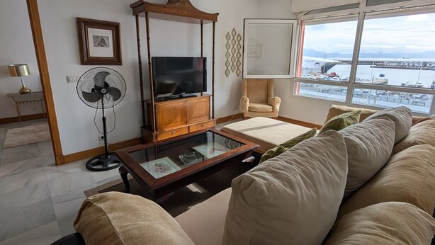 A living room with a large sofa, a TV on a wooden stand, a fan, and a view of a marina through large windows. - 293 - Apartamento El Puerto (Tarifa)