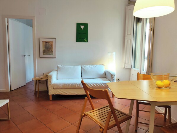 Une pièce avec un canapé blanc, une chaise en bois, une table sur laquelle se trouve un saladier rempli de citrons, un tableau encadré accroché au mur et un panneau vert.