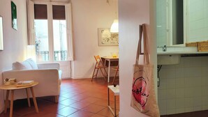 Una habitación con sofá, una mesa de comedor con sillas, una ventana con persianas, una lámpara fijada a la pared y una zona de cocina con fregadero.