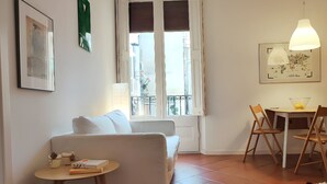 Un salón con un sofá blanco, una mesita redonda con libros, una zona de comedor con mesa y sillas, y un ventanal con vistas a edificios.