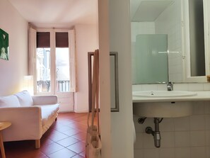 Une pièce comprenant un lit, une fenêtre avec des stores, une porte et une salle de bain avec un lavabo et un miroir.