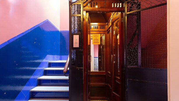 Una escalera de madera estrecha que conduce a un ascensor, con una pared revestida de azulejos azules en un lado y una pared revestida de azulejos rojos en el otro.