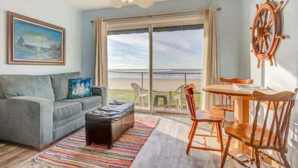 A living room with a sofa, ottoman, and a dining table with chairs. There is a large window with a view of the outdoors.