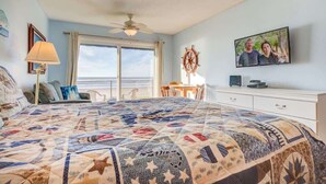 A bedroom with a large bed, a ceiling fan, a TV, a desk, and a view of the outdoors.