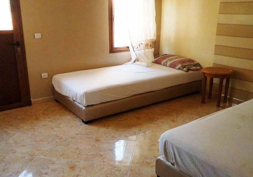 Una habitación pequeña con cama individual, suelo de baldosas, una ventana con cortinas y un taburete de madera.