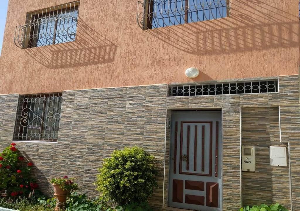 A building with a textured facade, a gated entrance, and a small garden with red flowers.