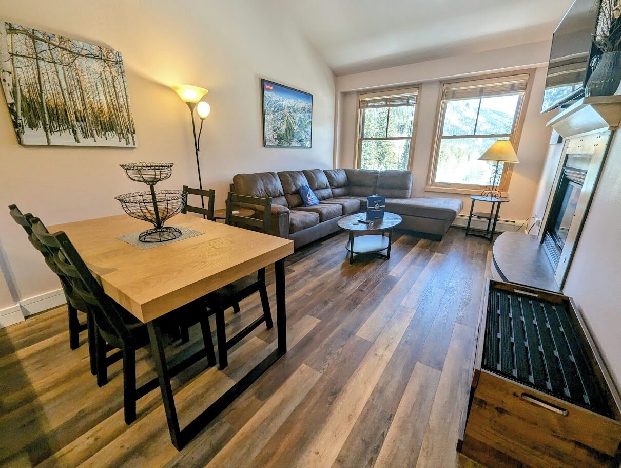 A spacious living room with a wooden dining table, a sofa, and a coffee table. There is a fireplace, a large window, and artwork on the walls.