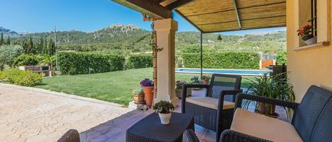A covered patio with wicker furniture and a view of a pool and greenery.