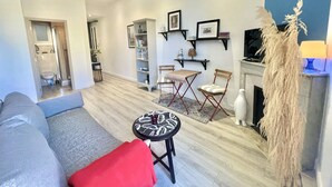 A modern living room with a grey sofa, red throw pillow, and a small round table. There is a fireplace with a decorative plant and a television mounted on the wall.