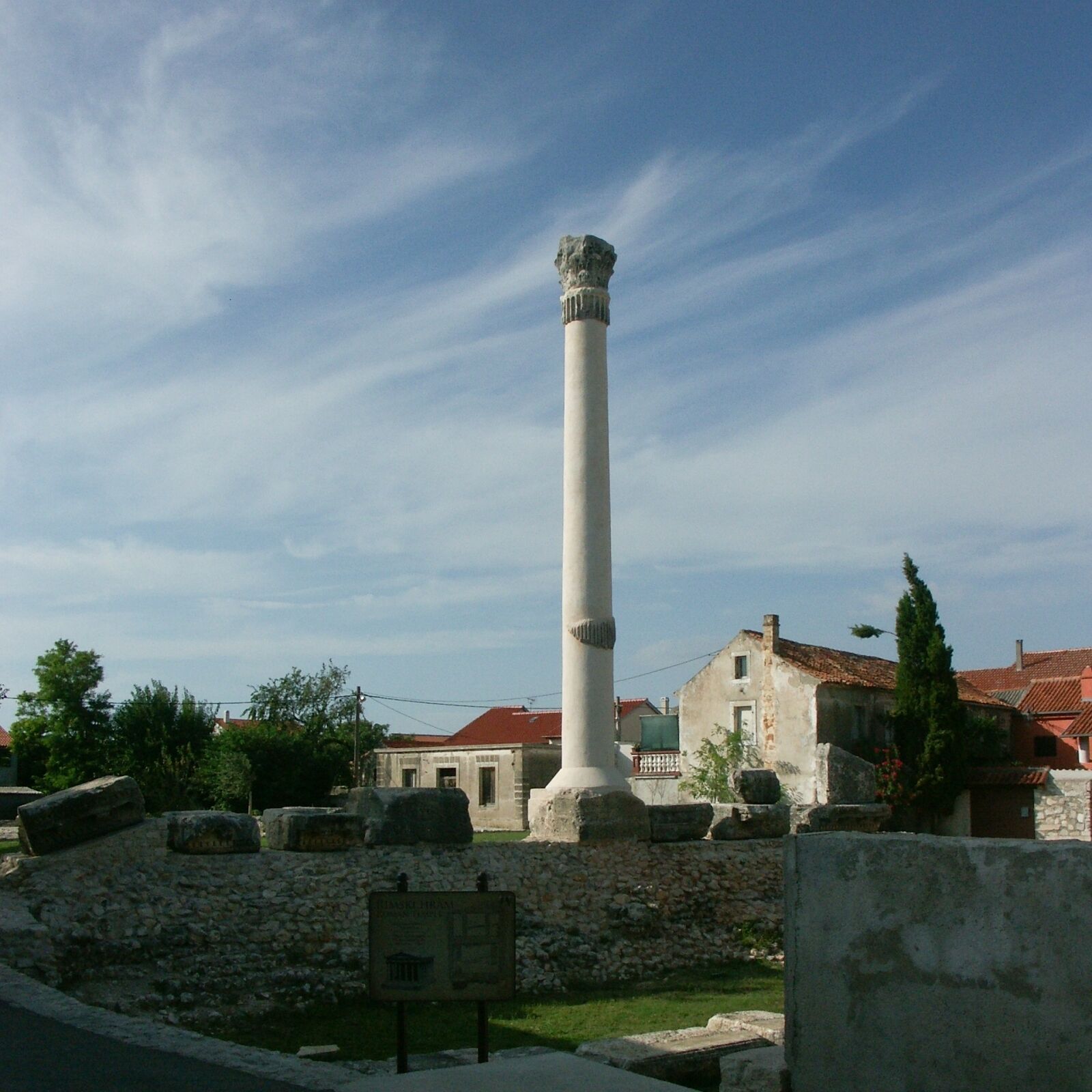 A tall white column with a capital at the top, surrounded by stone ruins and a stone wall.