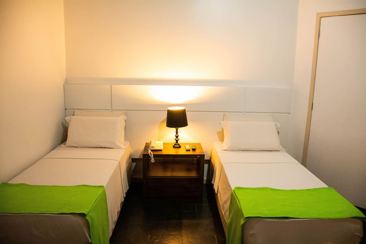 Quarto pequeno e bem iluminado com duas camas de solteiro, mesa de cabeceira com abajur e telefone.
