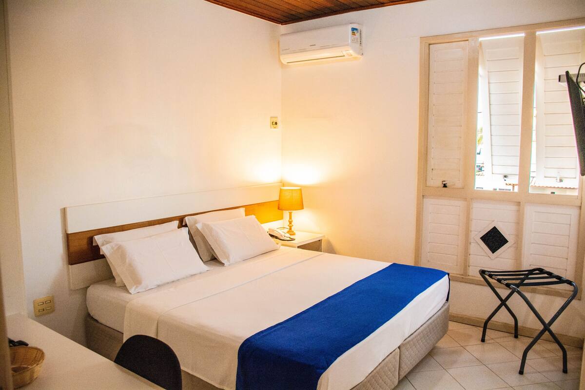 Quarto de hotel com cama, mesa de cabeceira com abajur, janela com persianas e ar condicionado.