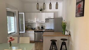 A modern kitchen with white cabinets, a dishwasher, a stove, and a coffee maker. There are two bar stools and a glass dining table with a vase of flowers. - Apartment in Bibinje with Terrace, Air condition, WIFI, Washing machine (877-3) (Bibinje)