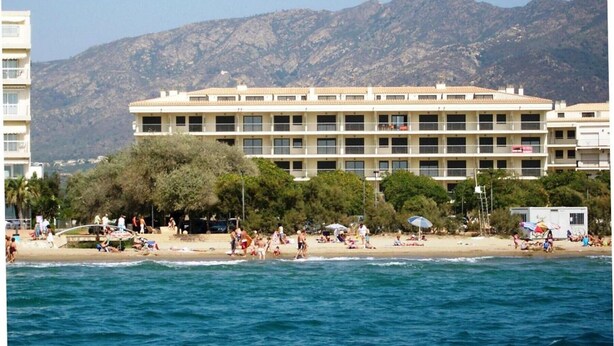 A beachfront hotel with multiple floors, a sandy beach, and mountains in the background. - MILENI 3 1 1 4 Roses (Rosas / Roses)