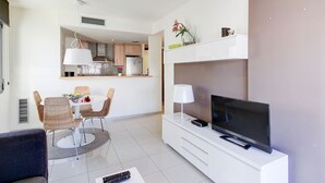 A modern living room with a flat-screen TV, a dining area with wicker chairs, and a kitchen in the background. - MILENI 3 1 1 4 Roses (Rosas / Roses)