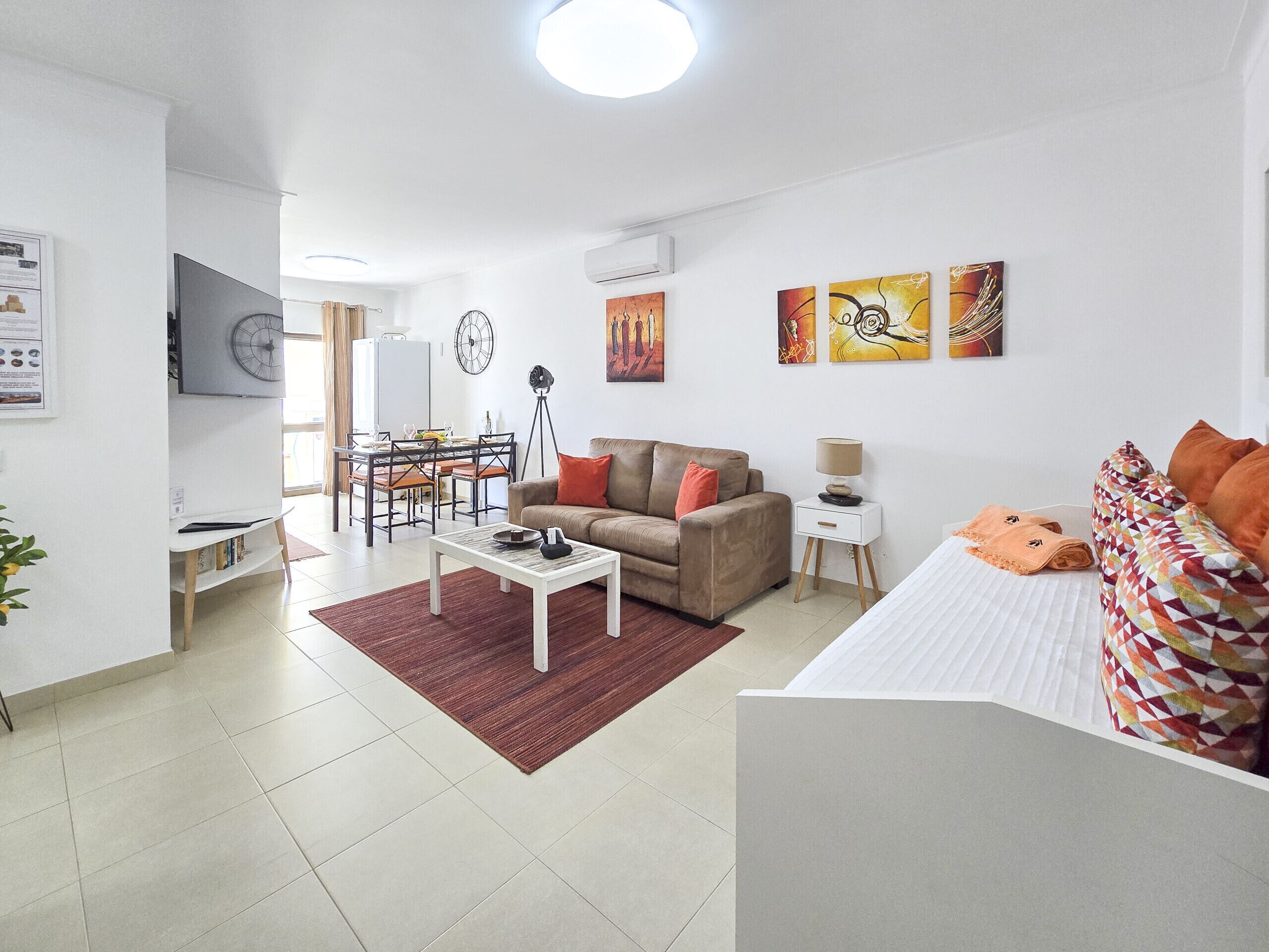 Ein modernes Wohnzimmer mit Sofa, Esstisch und Fernseher. Ein kleiner Beistelltisch mit Lampe sowie ein Bett mit gemusterten Kissen.