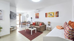 Ein modernes Wohnzimmer mit Sofa, Esstisch und Fernseher. Ein kleiner Beistelltisch mit Lampe sowie ein Bett mit gemusterten Kissen.
