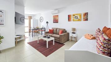 Una sala moderna con sofá, mesa de comedor y televisor. Hay una mesita auxiliar con lámpara y una cama con almohadones estampados.