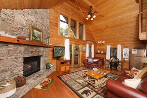 A cozy log cabin interior with a stone fireplace, wooden walls, a dining area, and a living room furnished with leather sofas and a coffee table.