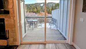 Una puerta corrediza de vidrio da a un patio exterior con una mesa y sillas, una chimenea de piedra y vistas a un área boscosa.