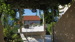 Un espace extérieur abrité, avec une table et des chaises, entouré de verdure et, au fond, un bâtiment dont le toit est recouvert de tuiles rouges.