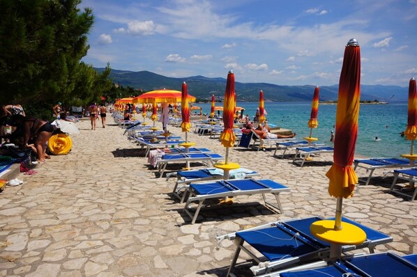 A beach with rows of sun loungers and umbrellas, a stone-paved walkway, and a view of the sea and mountains in the background. - Apartment in Povile with Terrace, Air condition, WIFI, Washing machine (3542-1) (Povile)