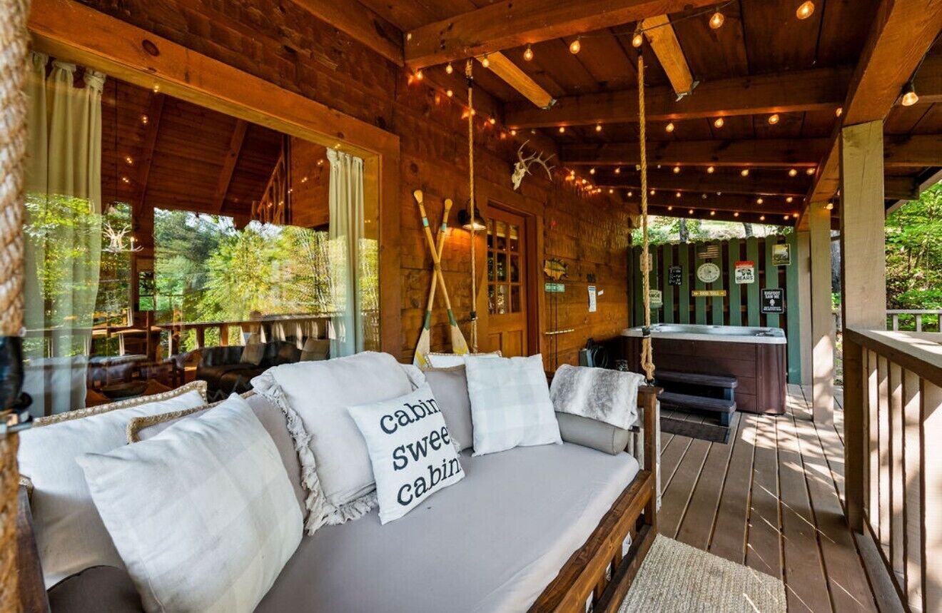 A cabin with a wooden deck, a sofa with pillows, a hot tub, and a view of the forest.
