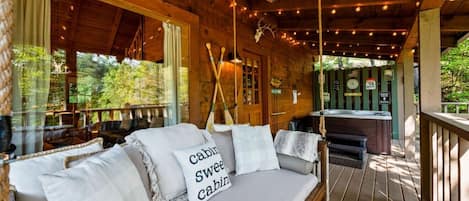 A cabin with a wooden deck, a sofa with pillows, a hot tub, and a view of the forest.