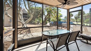 En overdækket terrasse med et glasbord og stole, store vinduer og en ventilator i loftet.