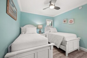 Une chambre à coucher avec deux lits blancs, un ventilateur de plafond, une table de chevet avec une lampe et une fenêtre avec des stores.