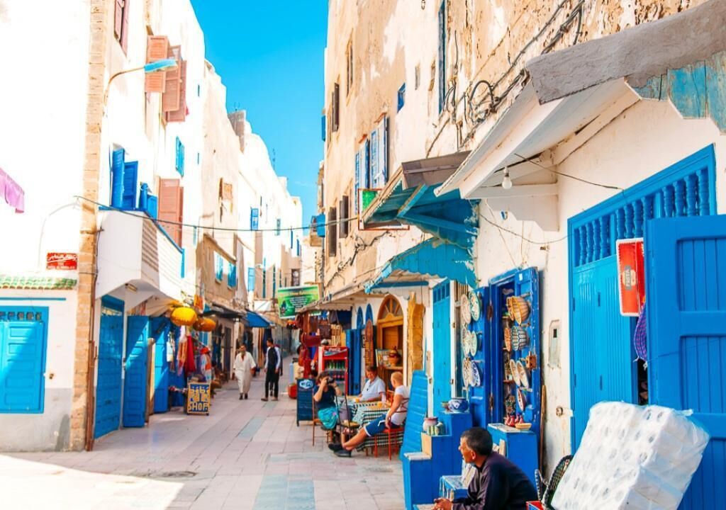 A narrow street with buildings on either side, featuring blue doors and shutters, and a few people sitting at outdoor tables.