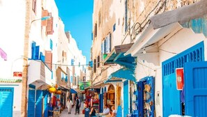 A narrow street with buildings on either side, featuring blue doors and shutters, and a few people sitting at outdoor tables. - Beautiful apartment in Essaouira 4158 (Essaouira)