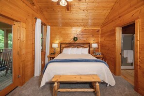 A wooden cabin bedroom with a bed, bedside lamps, a bench, and a view of the outdoors. - Hot Tub, Pool Table, Private Deck, Pool Access, Fishing, Putt Putt, Tennis (Sevierville)