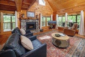 A log cabin interior with a stone fireplace, leather sofas, a round coffee table, and large windows.