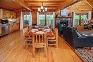 A wooden cabin interior with a dining table, chairs, a fireplace, a TV, and a stone fireplace surround.
