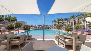 Un área junto a la piscina con sillones reclinables y mesas, una piscina azul cristalina y vistas a palmeras y edificios al fondo.