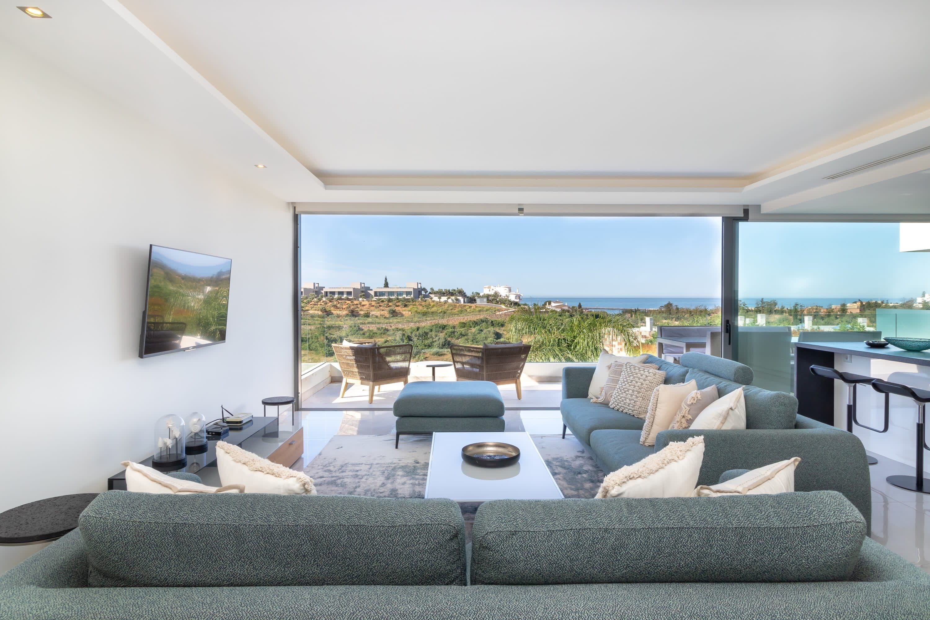 Una sala moderna con un sofá grande, una mesa de centro y un televisor montado en la pared. Se ve un paisaje costero a través de amplios ventanales.