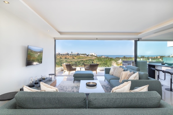 A modern living room with a large sofa, a coffee table, and a TV mounted on the wall. There is a view of a coastal landscape through large windows.