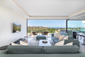 A modern living room with a large sofa, a coffee table, and a TV mounted on the wall. There is a view of a coastal landscape through large windows.