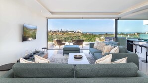Een moderne woonkamer met een grote bank, een bijzettafeltje en een aan de muur bevestigde televisie. Er is een balkon met uitzicht op zee en gebouwen.