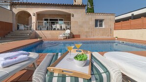 Un espace aménagé au bord de la piscine, avec des chaises longues, un plateau de boissons et un saladier de raisins, offrant une vue sur la piscine et le bâtiment.