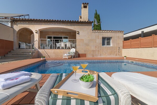 A poolside area with lounge chairs, a tray with drinks and a bowl of grapes, and a view of the pool and building.