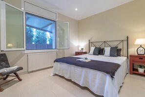 A bedroom with a large window, a bed with a white and blue bedspread, a bedside table with a lamp, and a chair.