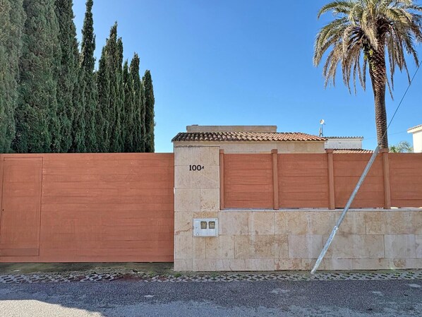 A gated property with a numbered gate, a palm tree, and tall evergreen trees in the background.