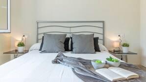 Un lit bien fait, recouvert d’une couverture grise, avec deux oreillers et une table de chevet sur laquelle se trouvent une lampe et une plante.