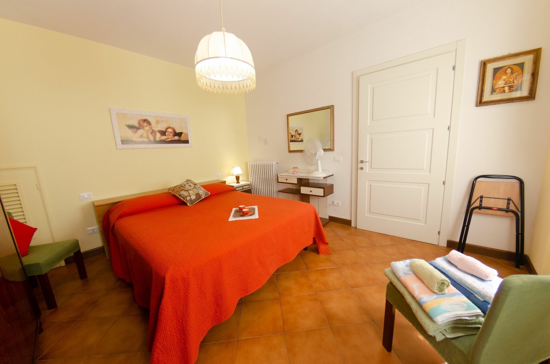 A bedroom with a large bed, a bedside table, a chair, a desk, a mirror, and a door.