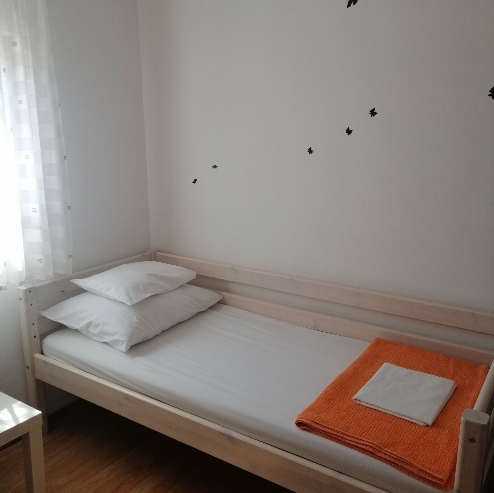 Ein Einzelbett mit weißer Bettwäsche und einem orangefarbenen Handtuch, ein kleiner Tisch und ein Fenster mit durchsichtigen Vorhängen.