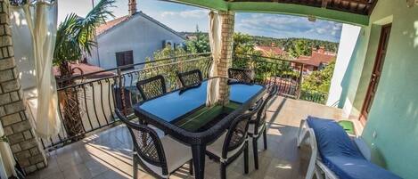 A covered outdoor dining area with a table and chairs, a lounge chair, and a view of the surrounding neighborhood.