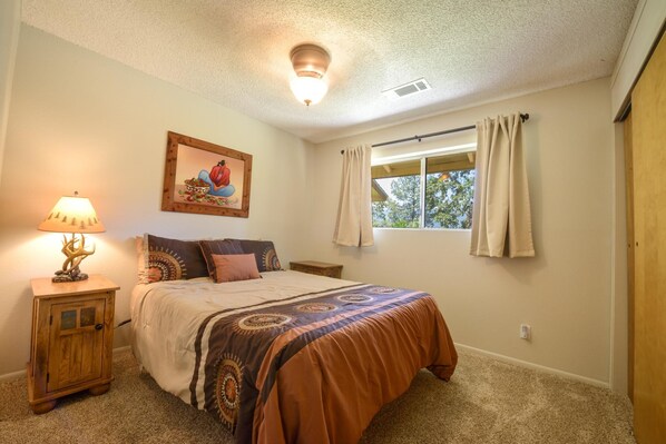 A bedroom with a bed, a nightstand, a lamp, a window with curtains, and a painting on the wall.